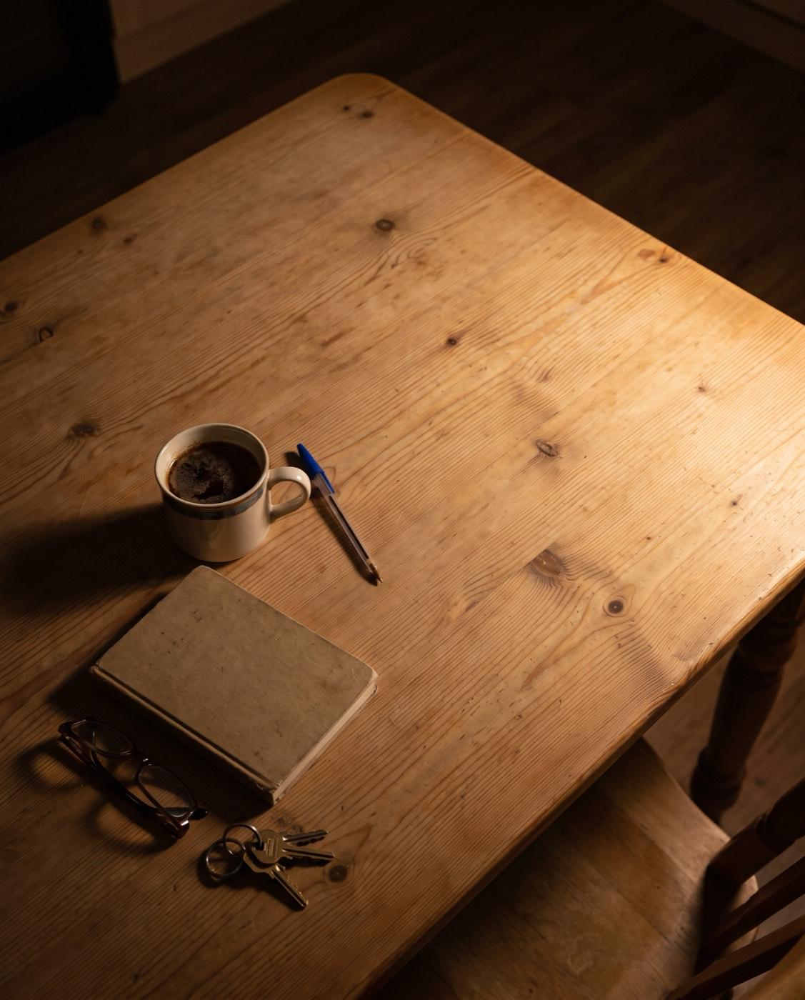 A kitchen table late at night used as a desk, seen from above: a mug of tea gone cold, a dropped pen, a closed notebook, reading glasses and keys.
